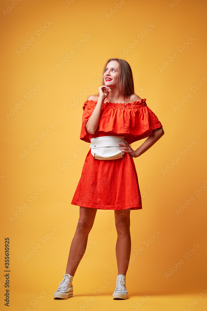 Summer urban fashion. Fun and colorful. Young pretty happy woman in orange dress posing against yellow background. Summer urban fashion. Fun and colorful. Young pretty happy woman in orange dress posing against yellow background.
