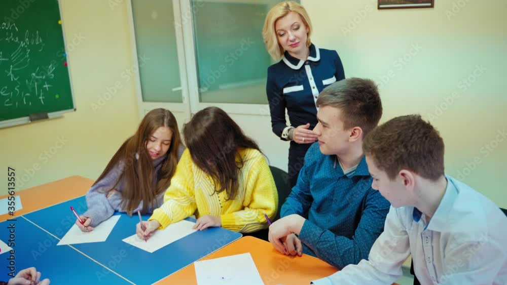 Female teacher talks with students. Teen pupils sitting in the classroom. Clever boy answering teacher questions at school. Female teacher talks with students. Teen pupils sitting in the classroom. Clever boy answering teacher questions at school.