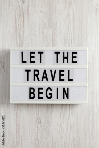 'Let the travel begin' on a lightbox on a white wooden background, top view. Flat lay, from above, overhead.