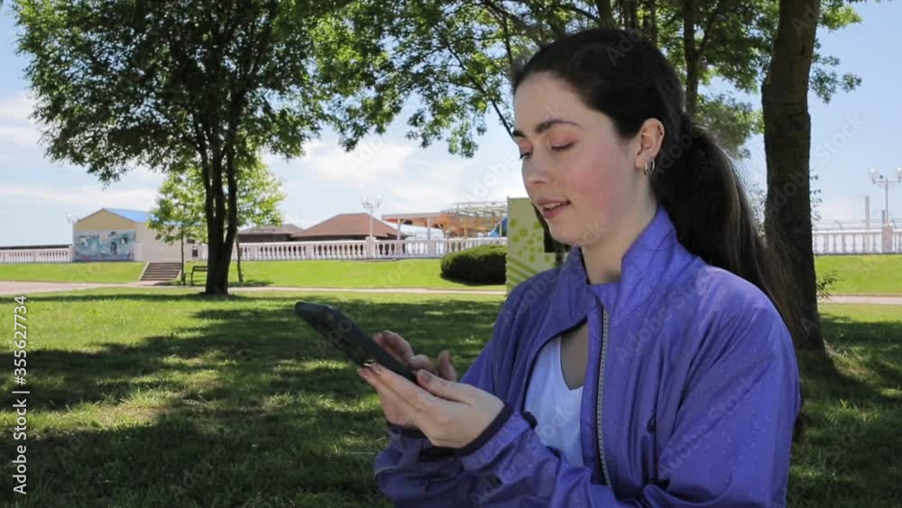 A young Caucasian woman is surfing the Internet on her phone. Park on the background. Concept of online communication and modern technologies A young Caucasian woman is surfing the Internet on her phone. Park on the background. Concept of online communication and modern technologies