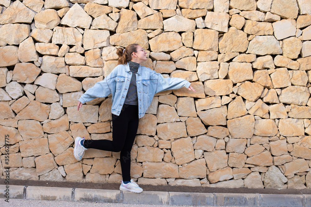 Young beautiful happy stylish hipster girl in denim jacket and glasses smiling having fun, teenager, stone wall background. Copy space