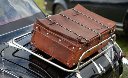 Close up of a vintage brown travel suitcase, strapped on a shiny chrome frame on the back of a black vintage car.  