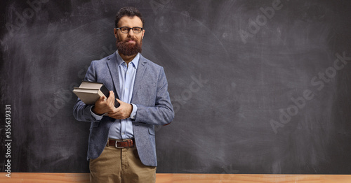 Bearded male teacher holding books and standing in front of a blackboard