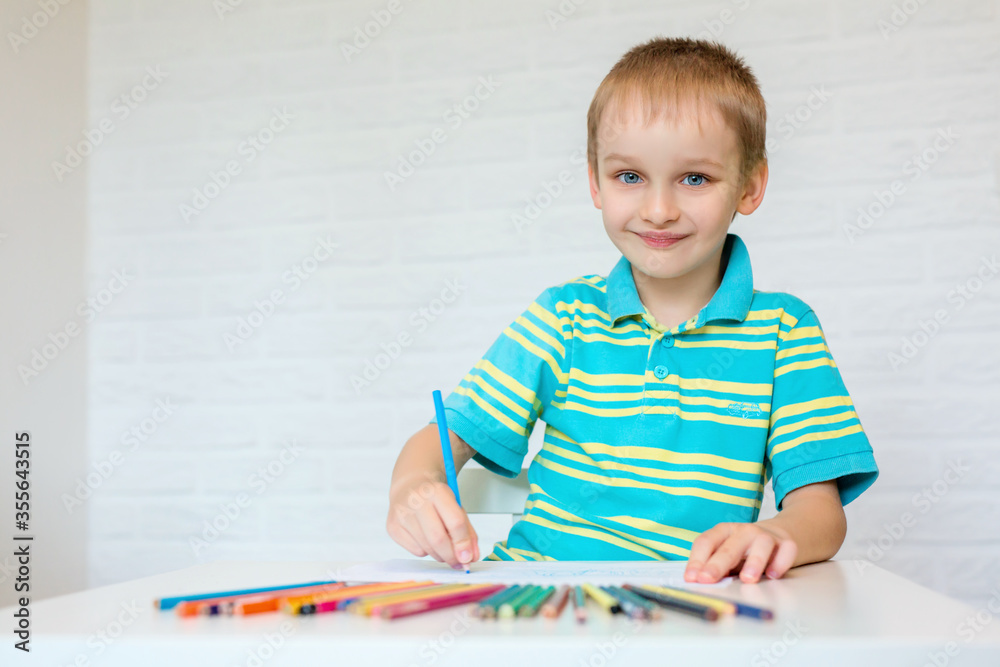 A beautiful boy draws a cute drawing with colored pencils. A cute child is sitting at a table and holding a pencil in his hand. Elementary school and homeschooling concept. A beautiful boy draws a cute drawing with colored pencils. A cute child is sitting at a table and holding a pencil in his hand. Elementary school and homeschooling concept.
