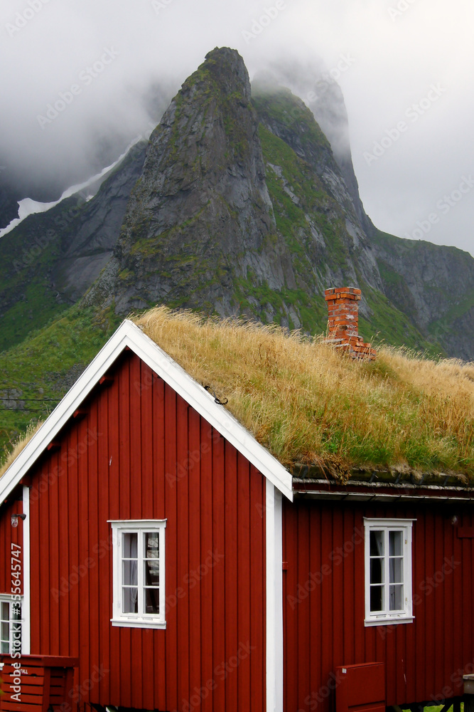 Reine, Lofoten Islands, Norway. Vertical view of red house with traditional grass roof in Reine, with mountain in mist in the background.