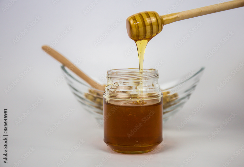 Glass jar with honey and a wooden stick on a white background. Pouring aromatic honey into jar, closeup. Glass jar with honey and a wooden stick on a white background. Pouring aromatic honey into jar, closeup.
