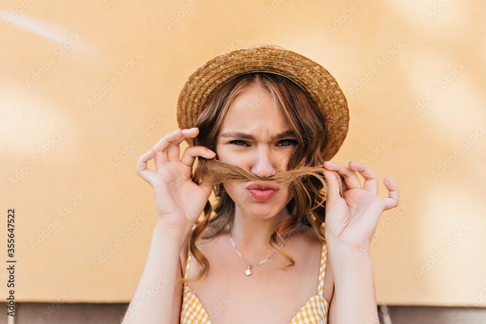 Funny dark-haired girl in hat making faces on yellow background. Joyful caucasian lady in summer attire posing in weekend.