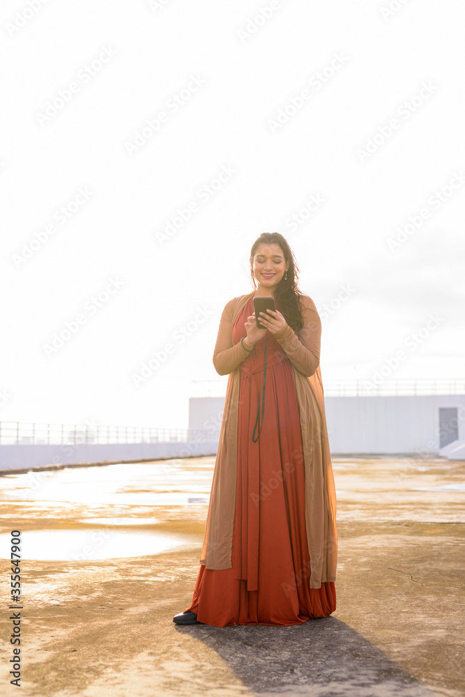 Full body shot of happy young beautiful Indian woman using phone at rooftop of the building Full body shot of happy young beautiful Indian woman using phone at rooftop of the building