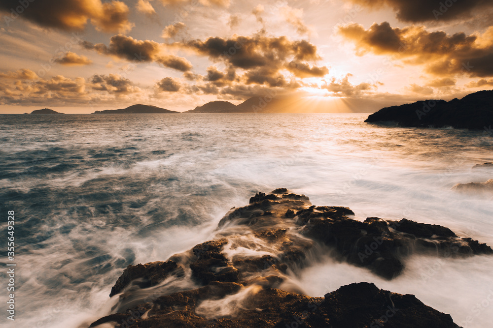 Obraz premium Mare mosso in tempesta, con nuvole, scogli, scoglier e sole al tramonto.
