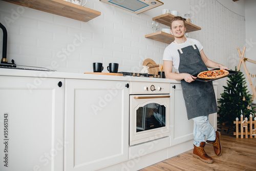 Homemade Italian pizza, handsome young male chef in dark apron holds baking sheet in bright kitchen. Cooking video blog concept