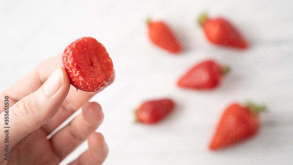 Fototapeta premium Hand holding bitten strawberry on white wooden background. Summer berry pleasure. Scattered strawberries