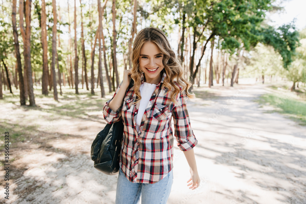 Confident european girl walking in park with backpack. Outdoor shot of romantic blonde lady wears checkered shirt in spring day. Confident european girl walking in park with backpack. Outdoor shot of romantic blonde lady wears checkered shirt in spring day.