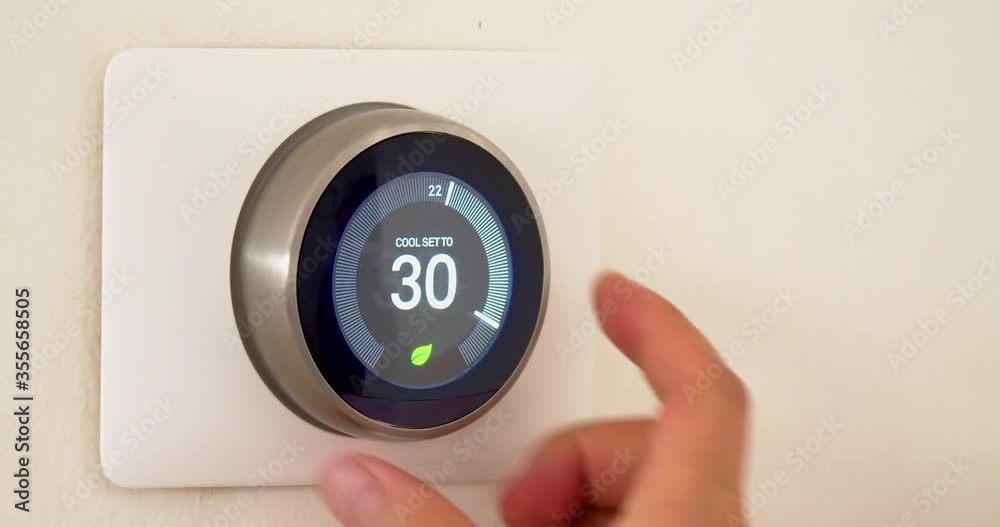 A persons hand saving energy during summer high temperatures on centigrade celsius metrics using a smart thermostat on a white wall. A persons hand saving energy during summer high temperatures on centigrade celsius metrics using a smart thermostat on a white wall.