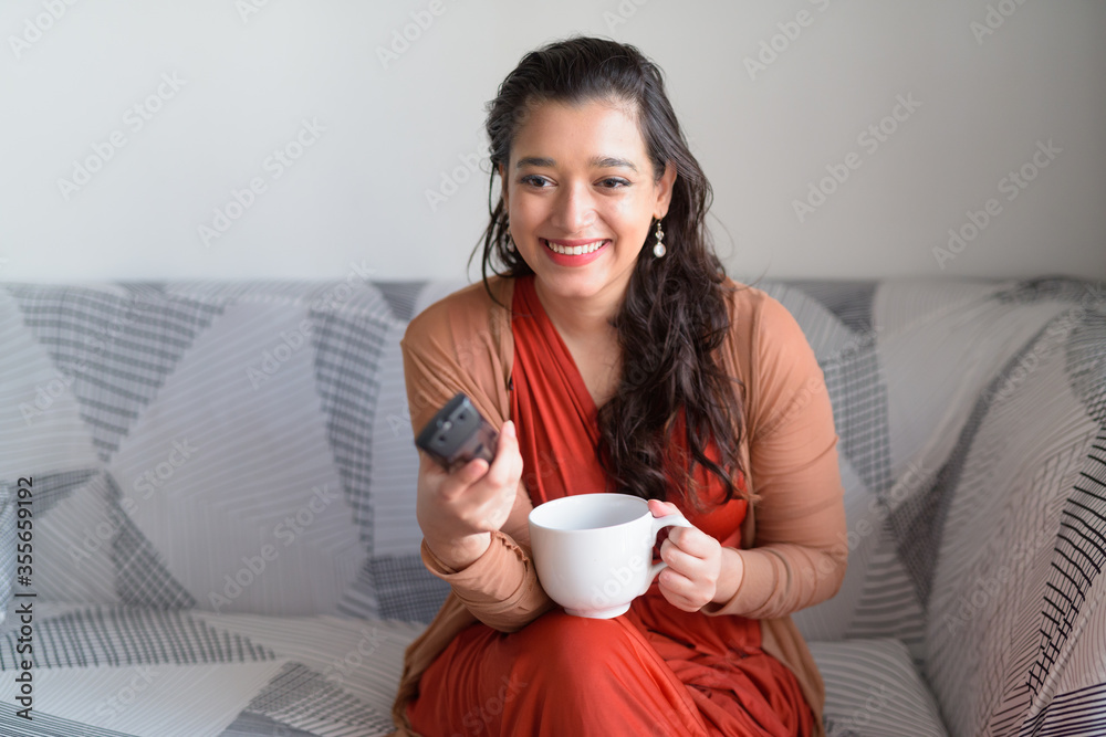 Happy young beautiful Indian woman with coffee laughing while watching tv at home