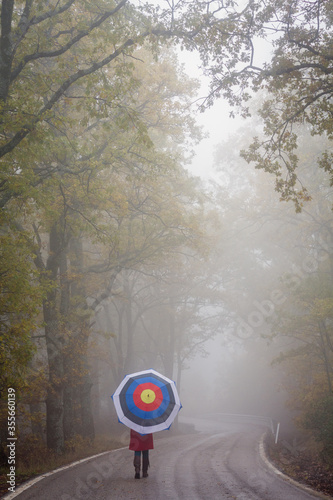 Girl with an archery shooting target umbrella walking on an empty road surrounded by scary autumn oak forest  in the fog.