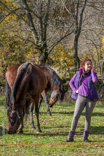 Girl talking on smart phone with two bay horses grazing in background.