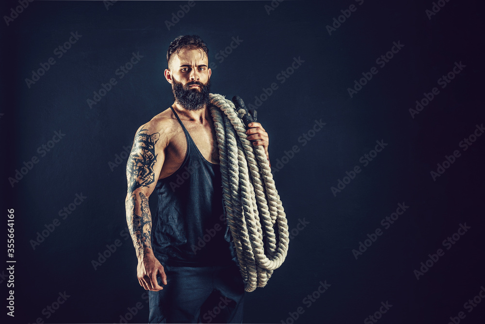 Bearded athletic looking bodybulder holding battle rope and fitness ball on dark studio background. Strength and motivation.