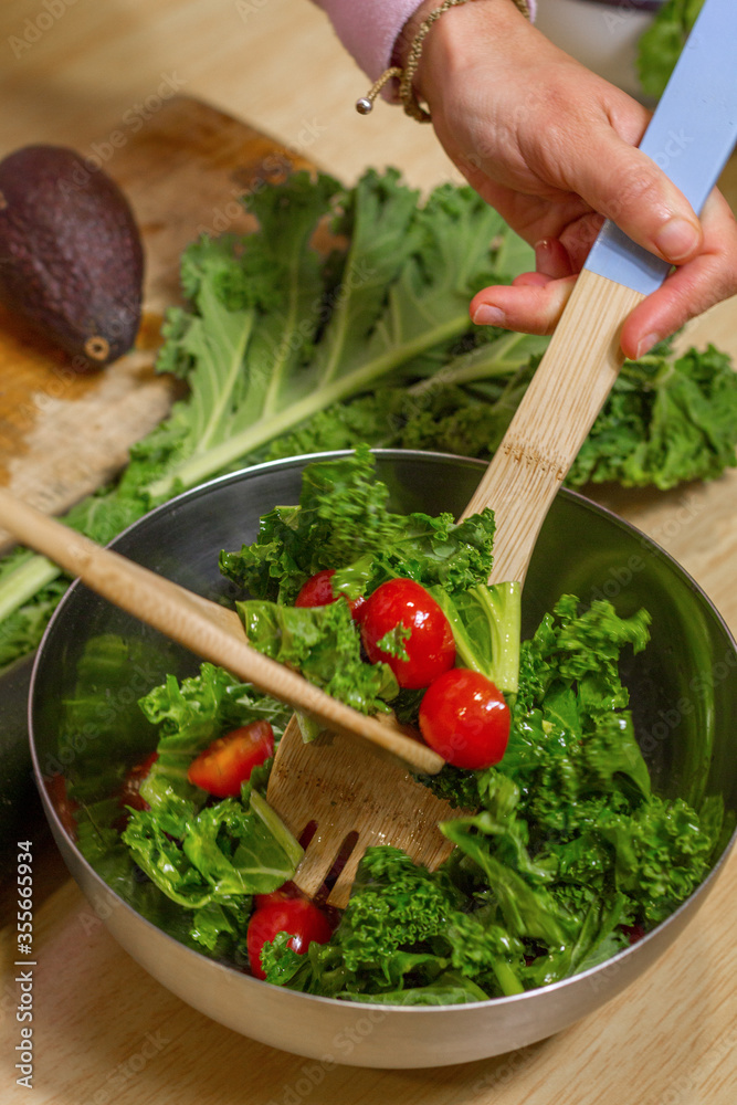 Close up of hands removing a kale and tomatoes salad in a metallic bowl with wooden spoons in a kitchen and an avocado in the background.