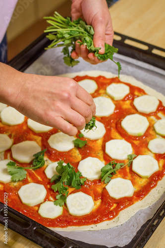 Female hands putting coriander leaves on a tomatoe base pizza with zucchini.