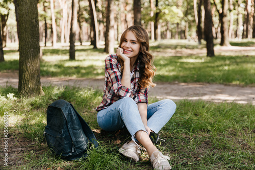 Charming female tourist posing in park with backpack. Outdoor shot of amazing curly girl sitting on the grass with smile.