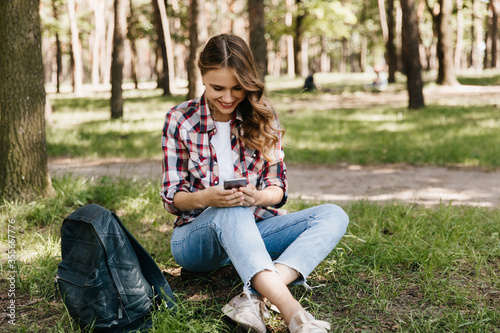 Pretty female traveler resting on the grass. Outdoor portrait of lovable girl in jeans posing in park with backpack.