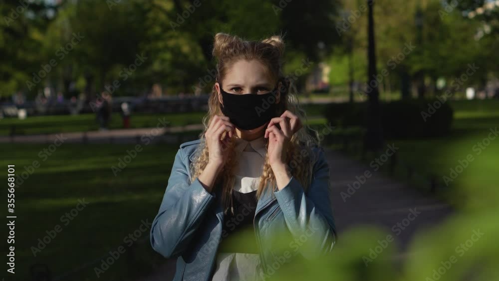 Young beautiful girl with luxurious curly hair puts on a black face mask in a public place. The blonde is protected from the spread of the virus in the park on the street. Young beautiful girl with luxurious curly hair puts on a black face mask in a public place. The blonde is protected from the spread of the virus in the park on the street.
