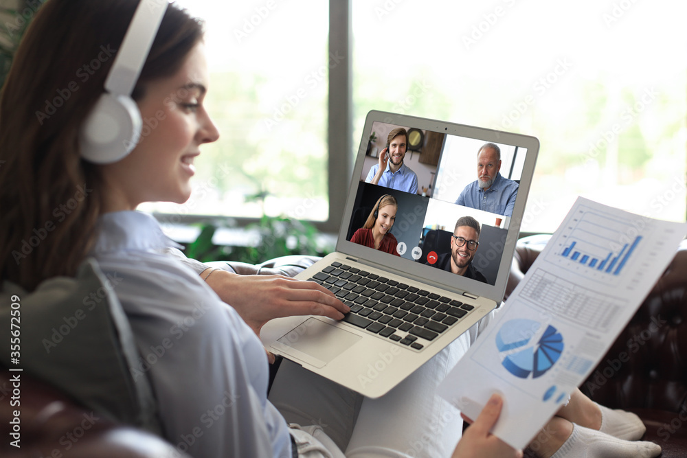 Business woman in headphones lying on sofa speak talking to her colleagues in video conference. Business team working from home using laptop, discussing financial report of their company. Business woman in headphones lying on sofa speak talking to her colleagues in video conference. Business team working from home using laptop, discussing financial report of their company.