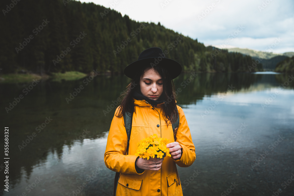 Φωτογραφία Thoughtful young woman wearing a hat, backpack and yellow jacket holding wild flowers bucket with lake and forest in the background