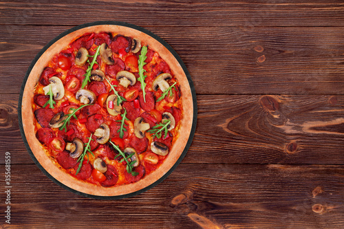 Delicious pizza hot with salami, arugula, cherry tomatoes, mushrooms and texas spice mix, on a round slate platter which is on wooden table, top view and copy space