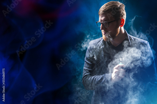 Vape. Vaper in the clouds of smoke. Human with a vape device in his hands. Man with a vape device on a dark background. Vaper with an electronic cigarette. Concept - sale of devices for a vaper.