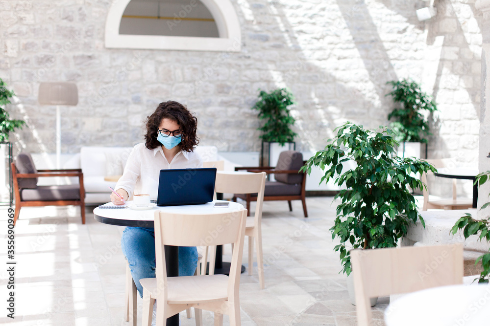 Fototapeta premium Young woman working safety in cafe indoors. Social distancing during quarantine. Freelancer wearing protective mask, using laptop. Comfortable office, coworking modern workplace with limited seating.