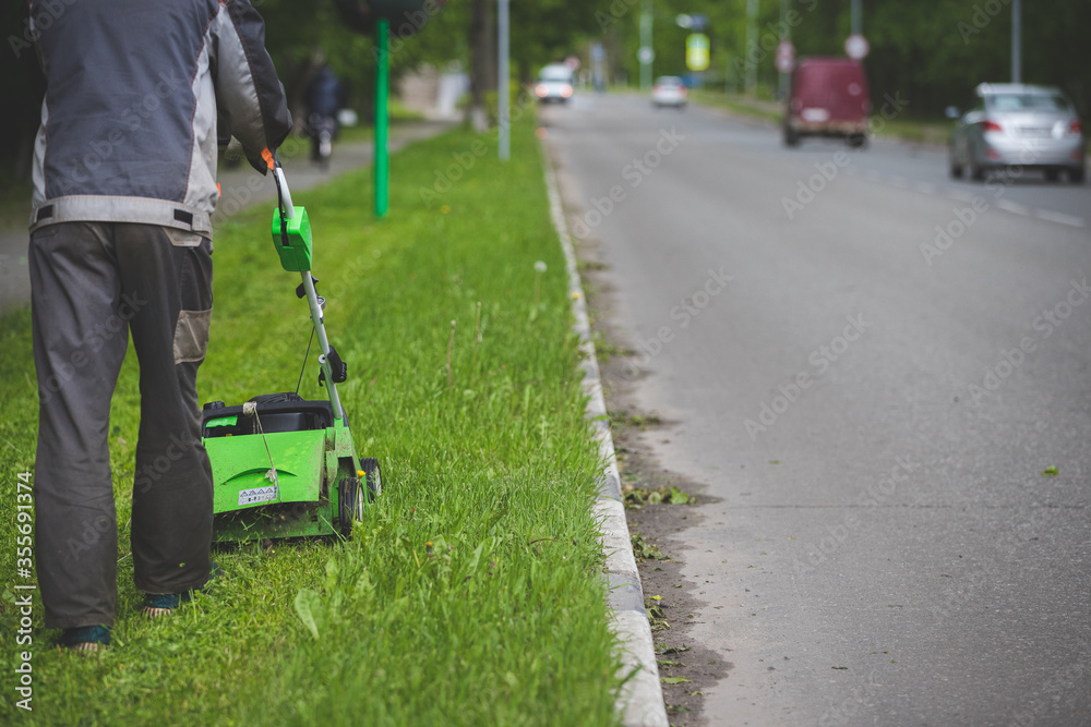 A worker in protective clothing and gloves with a gasoline mower on wheels walks along the lawn along the roadway. A man mows grass with dandelions next to passing cars in the city. Selective focus