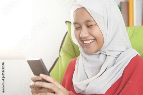Happy Asian muslim girl wearing hijab smiling while browsing trough internet or reading chat message on her phone