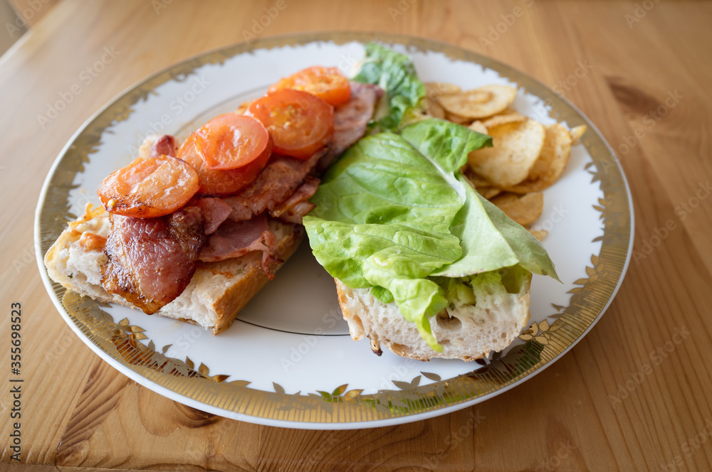 Homemade bacon and fried tomato and lettuce baguette open sandwich, Sometimes called a BLT before closing on a white plate with a gold trim with crisps. On a wooden table, lit with natural light Homemade bacon and fried tomato and lettuce baguette open sandwich, Sometimes called a BLT before closing on a white plate with a gold trim with crisps. On a wooden table, lit with natural light
