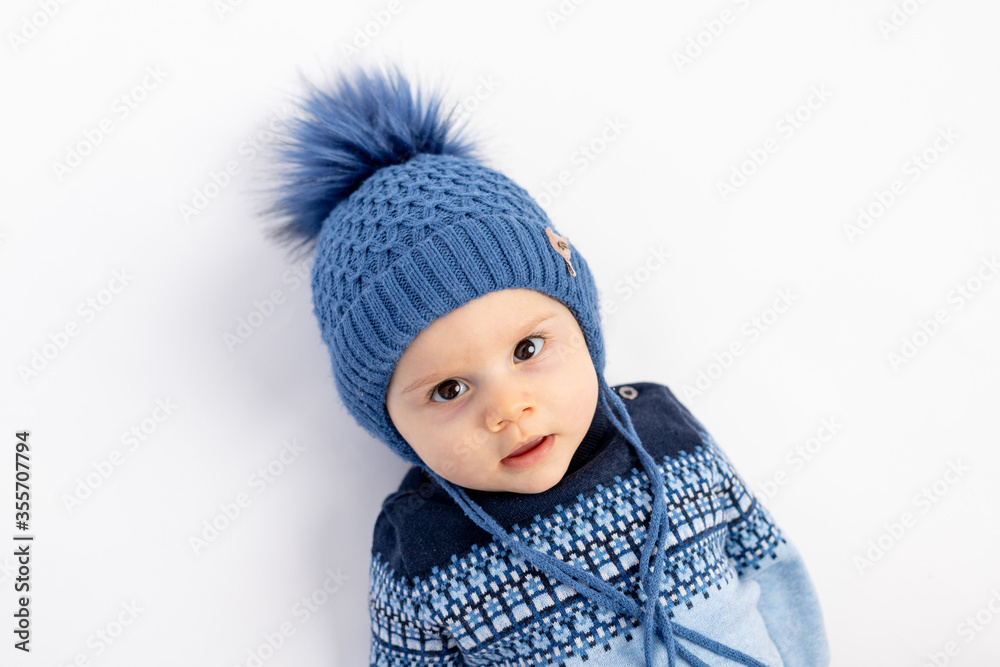 a smiling little boy in a Winter hat with a pompom lies and looks at the camera-a portrait of a 6-month-old child in warm winter clothes. a smiling little boy in a Winter hat with a pompom lies and looks at the camera-a portrait of a 6-month-old child in warm winter clothes.