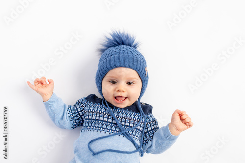 a smiling little boy in a Winter hat with a pompom lies and looks at the camera-a portrait of a 6-month-old child in warm winter clothes.
