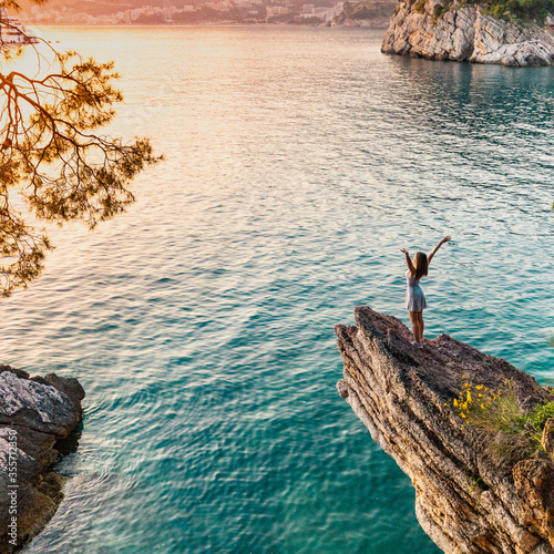 young woman stands on a ledge of a cliff above the sea on a background of sunset and a yacht