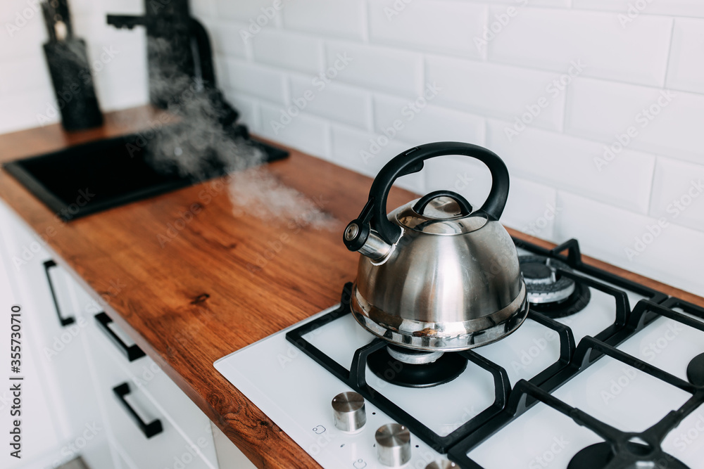 The kettle boils, steam from metal teapot on gas oven. Bright kitchen interior. White modern dining room. Wooden complete kitchen with gas oven.