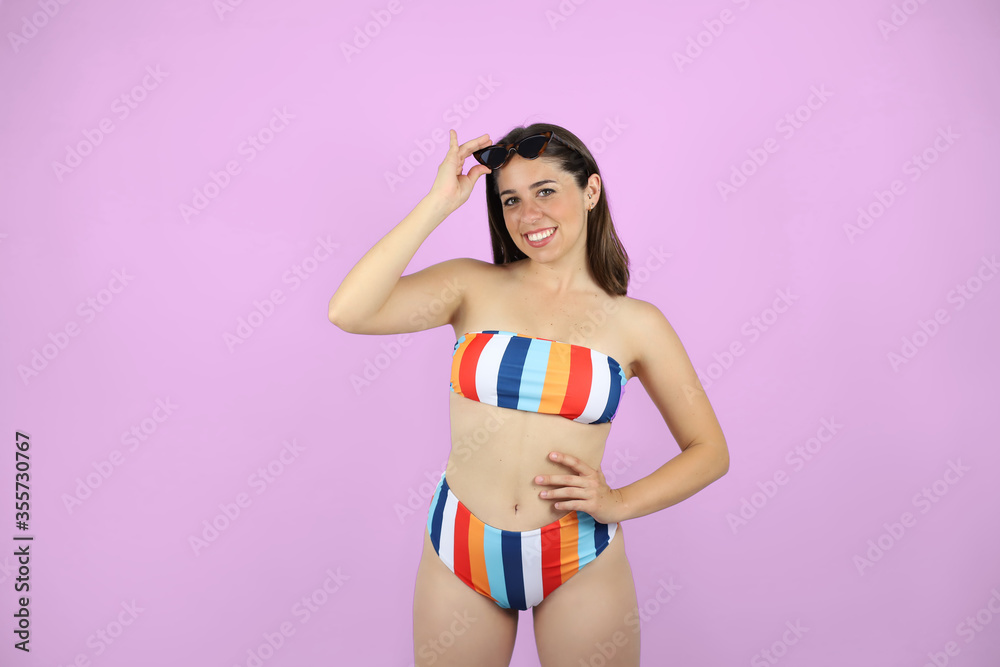Obraz premium Young beautiful woman standing over pink isolated background wearing bikini and sunglasses. very happy and holding the sunglasses with her hand