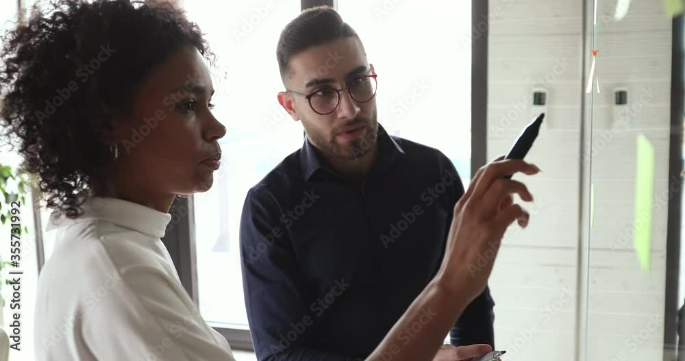 Two diverse team people brainstorm draw on glass wall discuss creative strategy at office meeting. African mentor coach teaching arab employee collaborating in research startup teamwork concept.