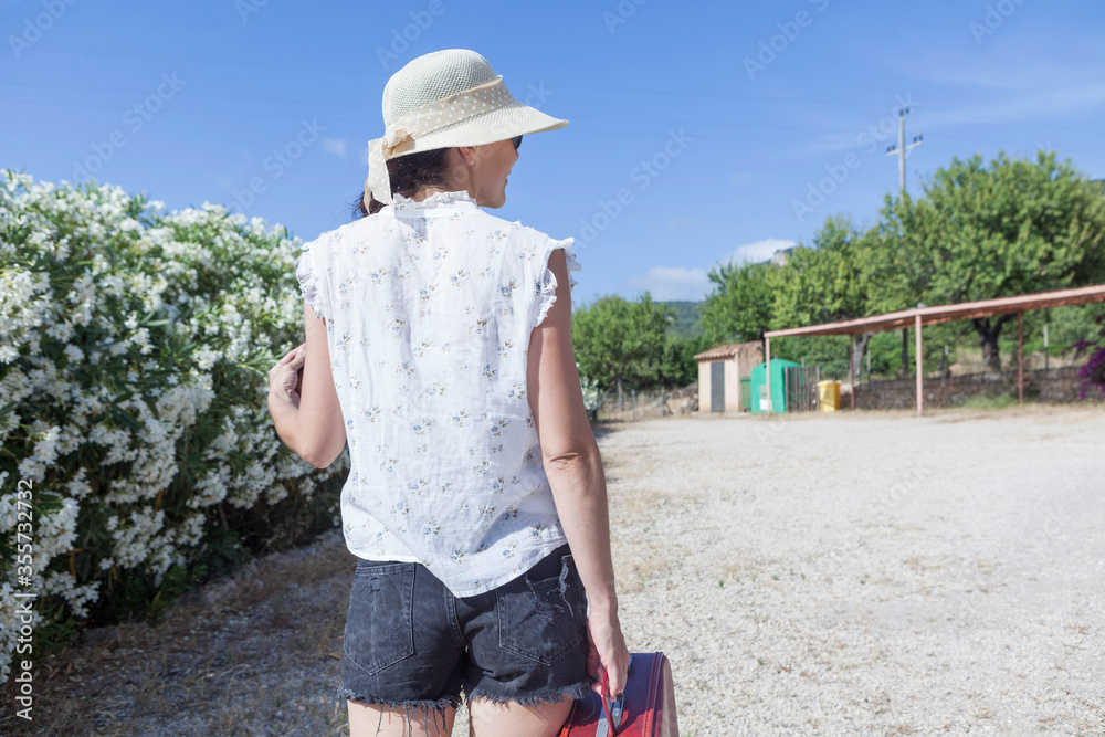 Wanderlust concept of  young woman walking on a way in spring summer wearing a hat and glasses holding a vintage red suitcase in her hand, her face expresses happiness and she is on vacation.