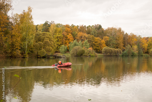 autumn landscape, trees green-yellow-red with a river and a red boat with a fisherman © Татьяна Ильиных