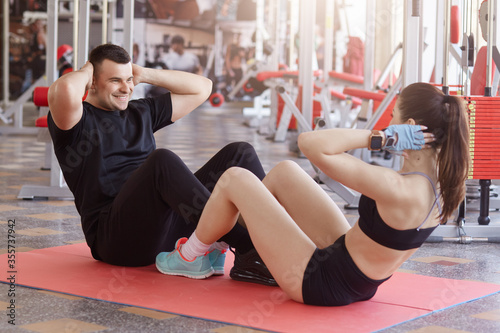 Young attractive Caucasian couple exercising together at gym. Fit man and woman sitting on floor in front of each other and doing abdominal exercises, keeping hands behind heads.