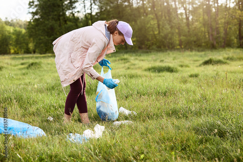 Woman being in middle of field between scattered cloth and holding garbage bag and picking trash during sunset. Environment pollution concept, collecting litter.