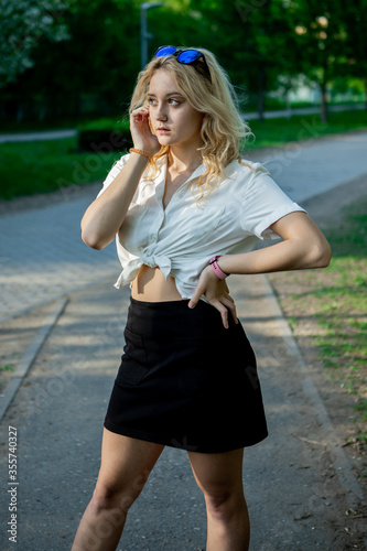 A teenage girl in a black skirt and white blouse. Light, elegant hair. Sunglasses. It stands in a Park on an asphalt path. © Алексей Дегтярев