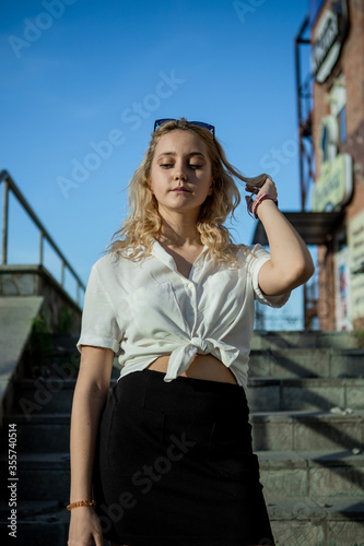 A young girl in a white shirt. She holds her blond hair in her hand. Blonde posing on the stairs on the street. Wearing sunglasses.