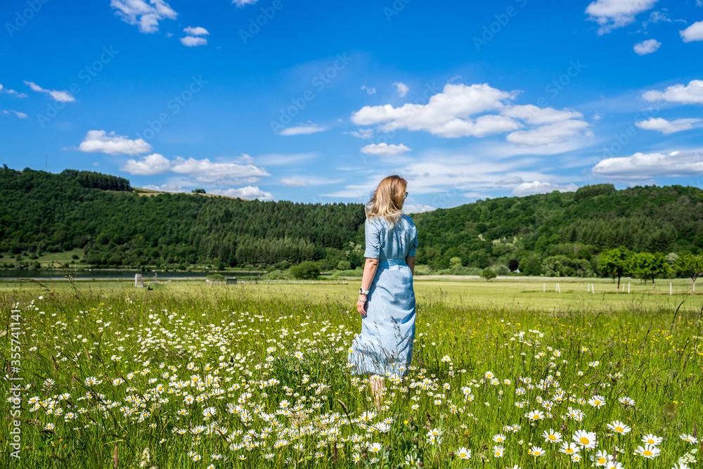 A happy girl in a light blue dress spinning and walking joyfully in a camomile field low angle shot against bright blue sky and white clouds. Germany, Eifel National Park , Meerfeld 