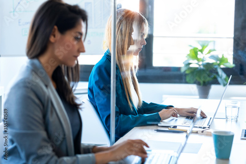 Two business women work with laptops on the partitioned desk in the coworking space. Concept of social distancing. photo