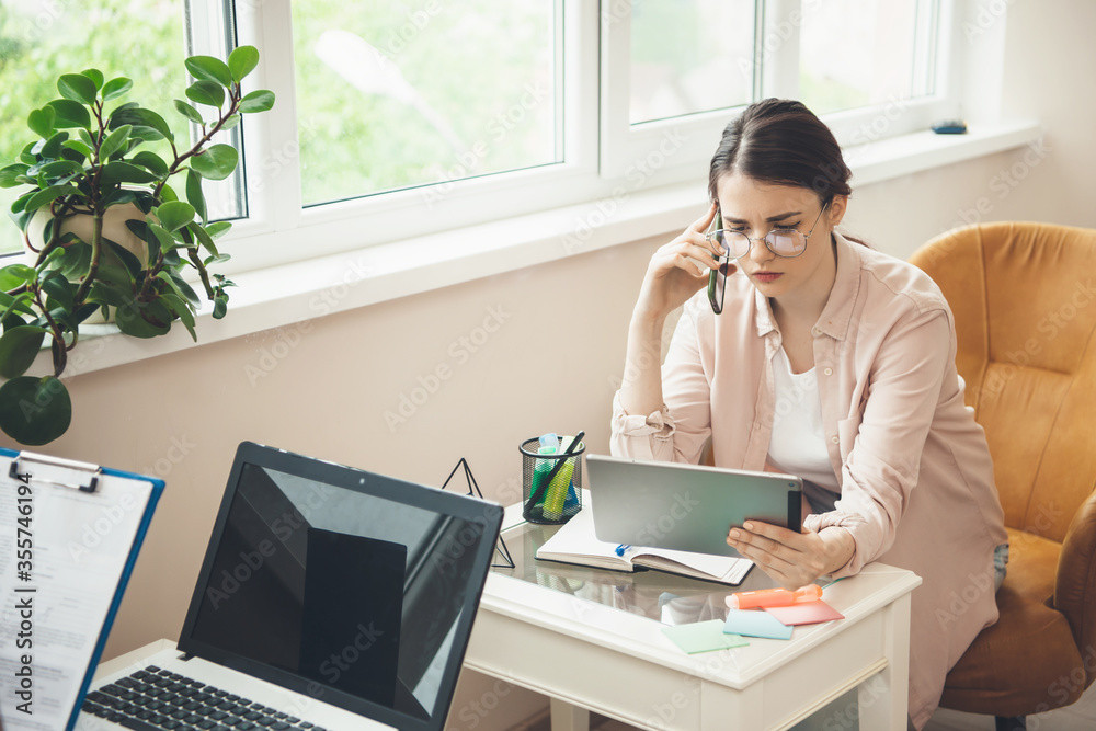 Young caucasian businesswoman talking on phone and working from home at the laptop and computer near the window Young caucasian businesswoman talking on phone and working from home at the laptop and computer near the window