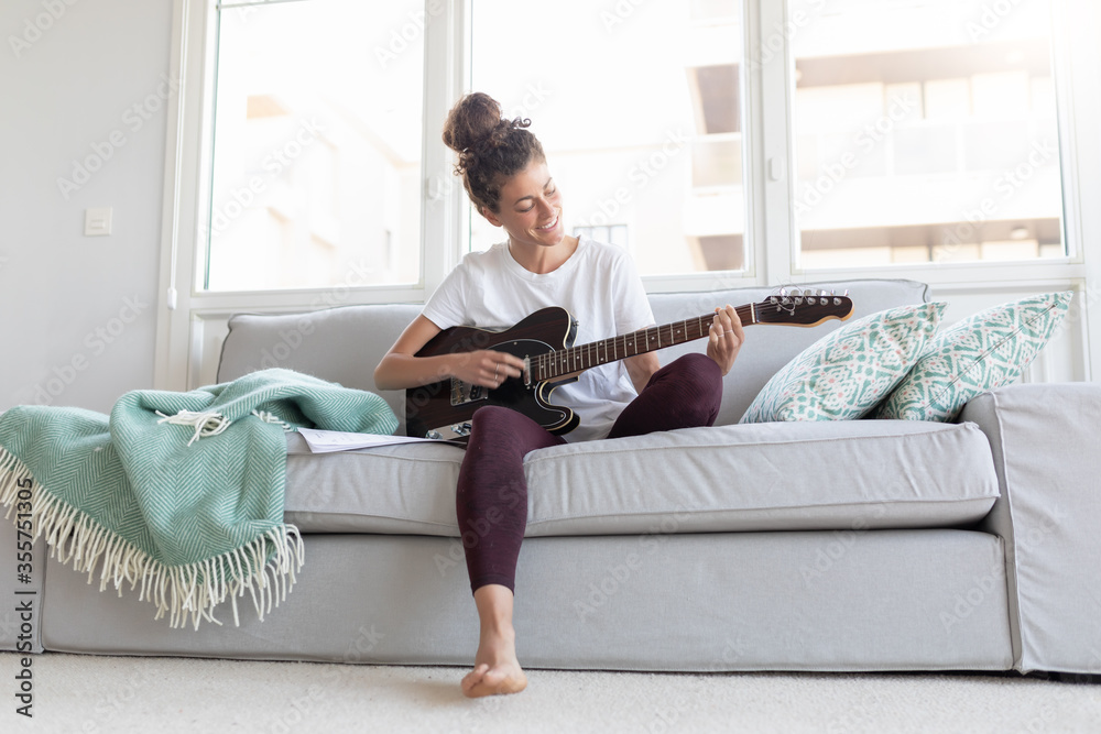 Woman sitting on an armchair at home learning to play the guitar in her spare time. Woman sitting on an armchair at home learning to play the guitar in her spare time.